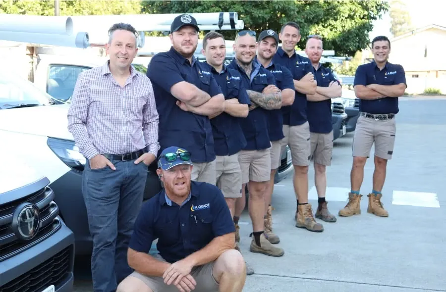 A Grade Plumbing team in navy uniforms grouped in front of their white work utes on a suburban street