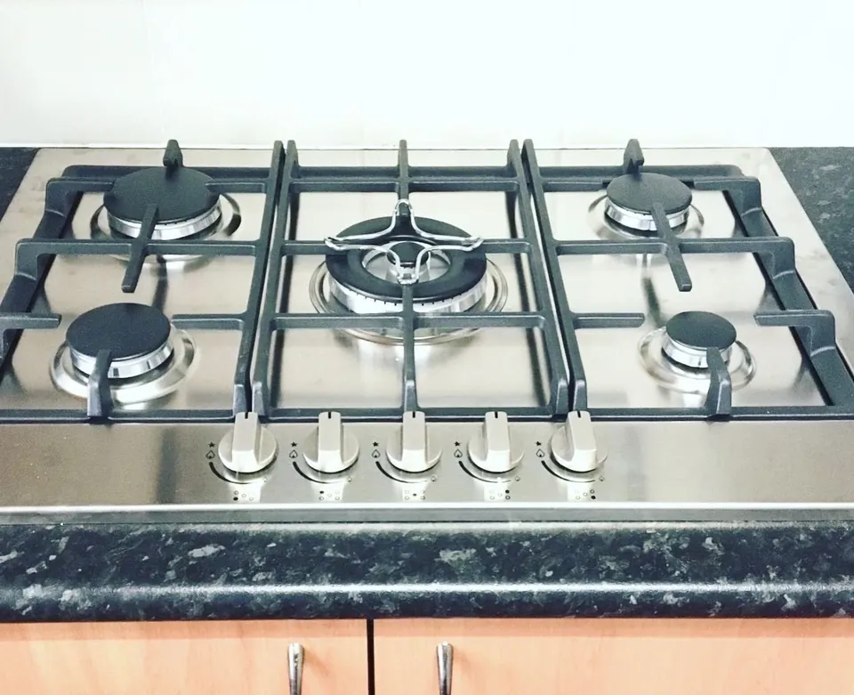 Five burner stainless steel gas cooktop installed into a dark stone benchtop above timber kitchen cabinetry